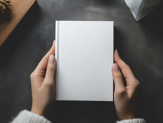 Elegant hands holding a blank white hardcover book on a dark moody background, minimalist reading concept with coffee and marble notebook for creative inspiration