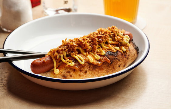 A hot dog is generously topped with mustard and crispy onions, resting in a white dish. Nearby, a cold drink sits, enhancing the inviting atmosphere of casual dining