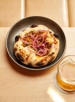 A circular flatbread is presented on a dark plate, adorned with slices of pickled onions and accompanied by a cold drink in a glass. The warm wooden table adds a cozy touch