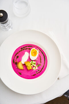 A vibrant beetroot soup is presented on a white plate, garnished with a halved egg and diced vegetables. The elegant setting enhances the dish's appeal