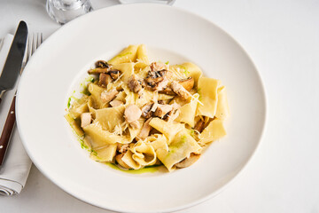 A hearty plate of pasta featuring tender chicken pieces and mushrooms is beautifully presented on a white dish. The meal is set on a table with elegant utensils, suggesting a fine dining experience
