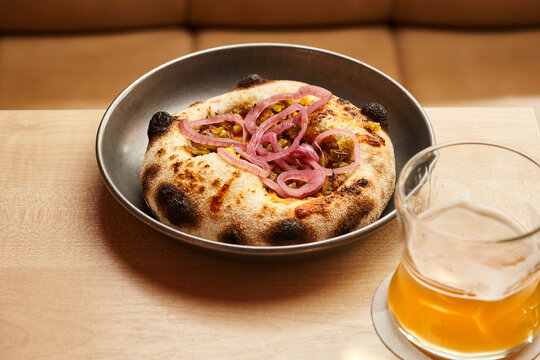A round flatbread sits on a metal plate, topped with thin slices of caramelized onions. Next to it, a tall glass of golden beer rests on a table in a warm, inviting atmosphere