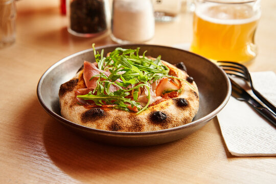 A flavorful flatbread rests on a metal plate, adorned with vibrant greens and thinly sliced cured meat. The warm atmosphere invites patrons to enjoy a casual meal