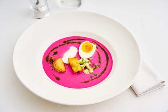 A vibrant bowl of beetroot soup features a perfectly boiled egg, diced vegetables, and a drizzle of sauce, all presented on a stylish white plate in an elegant setting