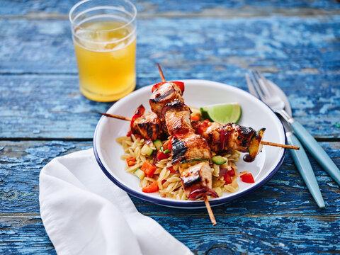 Succulent grilled chicken skewers are placed on a bed of orzo mixed with chopped vegetables. A chilled beverage complements the dish, all set on a rustic blue table
