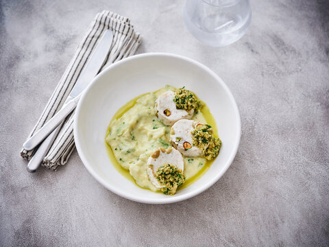 A plate features Pistachio-Stuffed Chicken Breasts resting on a smooth, creamy sauce drizzled with olive oil. This appetizing meal is set on a rustic surface with minimal tableware