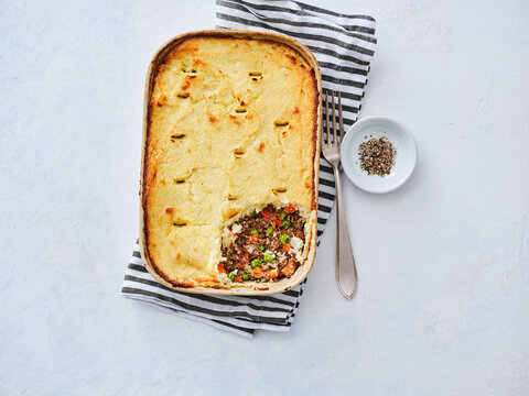 A hearty shepherd's pie sits in a ceramic dish, featuring a golden mashed potato topping and a rich filling. Accompanied by seasoning on a small plate, it invites a hearty meal