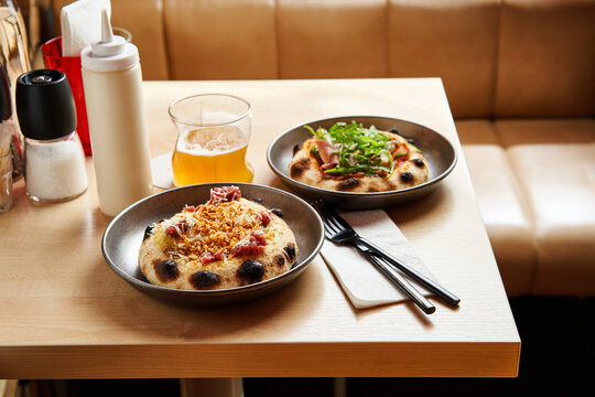 Two artisanal pinsa pizzas sit on a wooden table, topped with fresh greens and crispy elements. A cold craft beer accompanies the meal in a vibrant dining space, perfect for casual gatherings