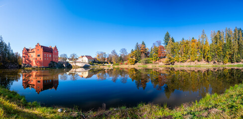 Fototapeta premium Panoramic view of Cervena Lhota Castle in the Czech Republic, surrounded by a lake reflecting its image. Autumn trees in vibrant colors and sunny weather create a picturesque, serene scene.
