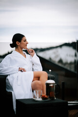 Woman in a white robe enjoys tea on a terrace with snow-capped mountains in the background during a peaceful morning