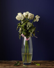 Wilted white roses in a glass vase with cloudy water on a wooden table, dark blue background, fallen leaves.