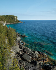 A lookout view of the deep blue and turquoise waters along a rocky shoreline, cliffs, and a cape point, surrounded by lush forests, and a far away island in the distance. 