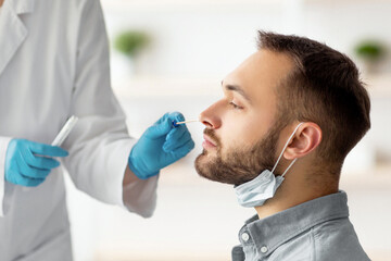 Naklejka premium Covid diagnostic procedure. Doctor making nasal PCR test for young Caucasian man, using sterile swab stick at home. Millennial guy undergoing coronavirus analysis, checking health