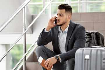 Worried Middle Eastern Man Talking On Cellphone While Sitting With Luggage On Stairs At Airport...