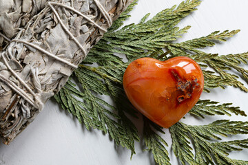 A top view image of a red carnelian crystal heart with white sage smudge stick wrapped with string and vintage metal key on a green cedar bow.