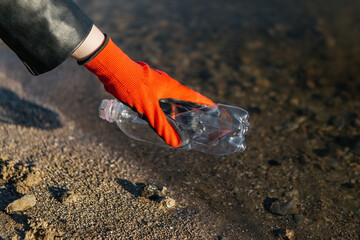 Close-up of a volunteer's hand wearing an orange glove picking up a plastic bottle from the edge of a polluted river, highlighting environmental pollution and cleanup efforts. Ecology, earth day