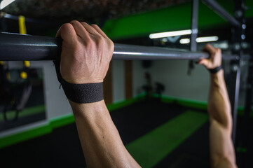 Close-up of strong hands gripping a pull-up bar with wrist straps in a modern gym. Strength...