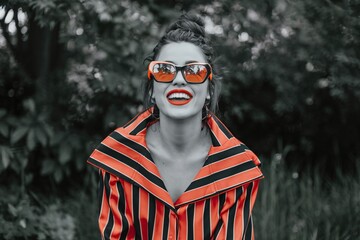 Vibrant Woman Laughing in Striped Shirt. A joyful woman wearing a bold striped shirt and orange sunglasses laughs heartily, exuding confidence and happiness against a lush green backdrop.