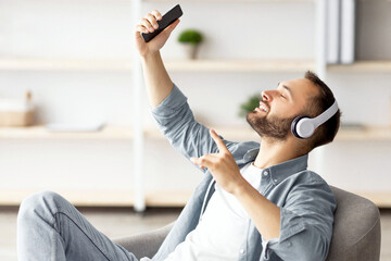 Smiling young Caucasian guy in headphones listening to music or audio book, sitting in armchair at home. Happy millennial man relaxing to his favorite song indoors. Weekend hobbies