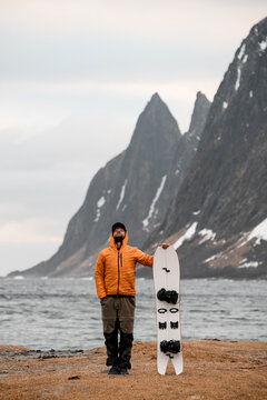 Young man in warm clothes is standing on the ocean shore, holding his splitboard