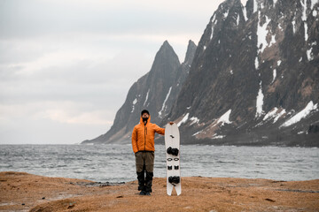 Young man in an orange jacket is standing on the shore holding his splitboard