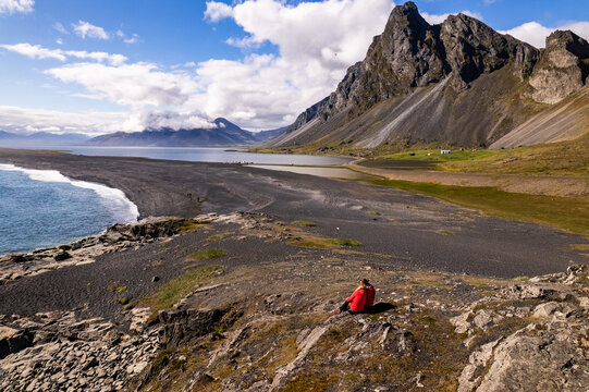Scenic aerial drone view of couple in red jackets in Hvalnes Nature Reserve Black Beach and amazing mountains in background by the Atlantic Ocean in Iceland