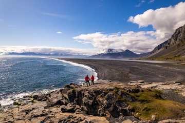 Scenic aerial drone view of couple in red jackets in Hvalnes Nature Reserve Black Beach and amazing mountains in background by the Atlantic Ocean in Iceland