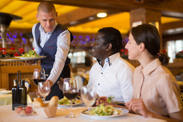 Waiter serving meals for positive interracial couple at restaurant, putting plates with food on table
