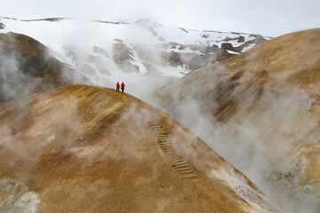 Scenic aerial drone view of a couple hikers in red jackets and foggy Kerlingarfjoll mountain path amidst desert and snowy terrain in Iceland