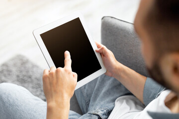 Advertisement concept. Millennial man using tablet computer with blank black screen, browsing website or demonstrating new app, sitting on couch at home. Mockup for your design