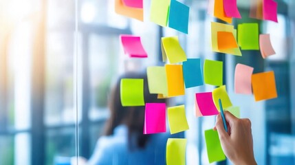 Brainstorming Session:  A  hand writes on a sticky note on a glass wall covered with colorful notes.