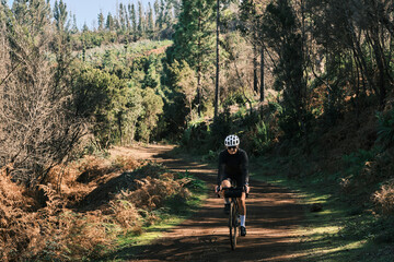 A cyclist rides a gravel bike on a scenic forest trail. Sunlight filters through the trees, casting long shadows. The cyclist is wearing a helmet and dark clothing. Tenerife, Canary Island