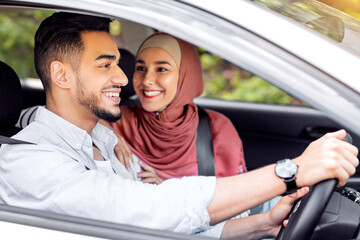 Smiling excited millennial middle eastern woman in hijab looks at male at steering wheel, have good news in car. Couple go to journey, road travel, vacation, love relationship and family vacation