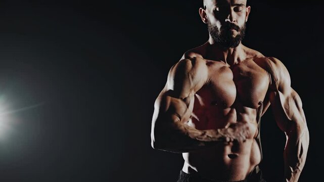 Muscular sportsman displaying well trained physique, flexing biceps and triceps under dramatic spotlight against dark background, highlighting muscle definition and strength