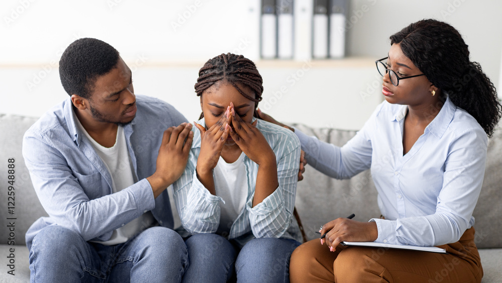 Wall mural Young African American woman crying at therapist's office, affectionate husband supporting her with professional psychologist. Millennial lady with her spouse on session with marital counselor