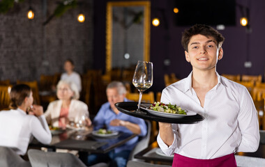 Young waiter guy is standing with order tray in spacious guest room in restaurant. Male restaurateur smiles and looks at camera. Cafe patrons eating in background.