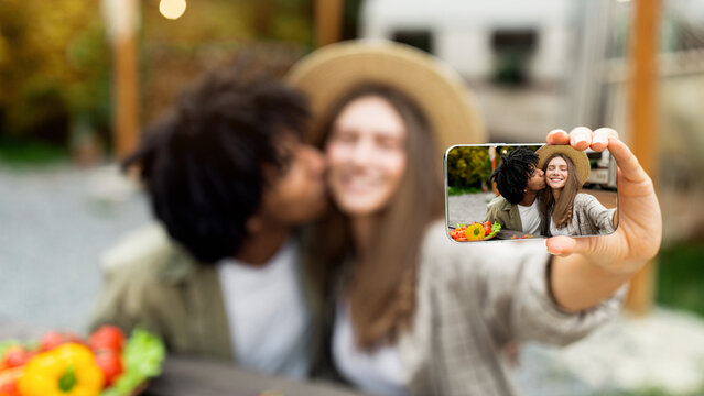 Affectionate African American guy kissing his Caucasian girlfriend, taking selfie on mobile phone near motorhome on camping trip in autumn, selective focus on mobile device with photo on screen