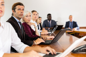 Focused mature white business man in formal suit sitting with colleagues in conference room and absorbedly listening to speaker's presentation during corporate group meeting