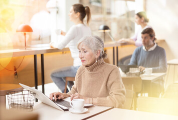 Obraz premium Positive European old woman drinking and enjoying coffee while working on laptop in cafe