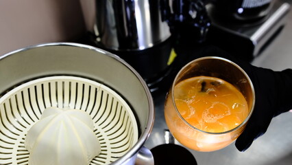 A glass of freshly squeezed citrus juice from ripe oranges served with ice in a clear glass placed next to a juicer held by a barista in a modern cafe setting emphasizing freshness and health