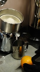 A barista in black disposable gloves slices and squeezes fresh oranges on a professional juicer into a metal pitcher in a modern kitchen preparing a refreshing citrus beverage with vitamin C