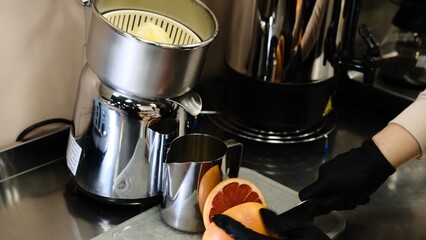 Barista in black disposable gloves cuts squeezes fresh grapefruit on professional juicer into a metal pitcher, preparing fresh juice in clean kitchen with focus on healthy eating and vibrant drinks.