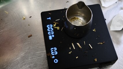 Weighing loose tea leaves in metal container on digital scales for portioning and brewing preparation close up high quality fresh tea leaves precise measurement traditional tea making process.