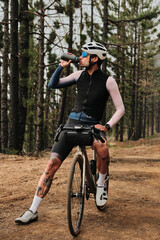 A cyclist takes a break, enjoying the scenery and hydrating during a ride through a pine forest.  He's wearing cycling gear and is sitting on his bike. 