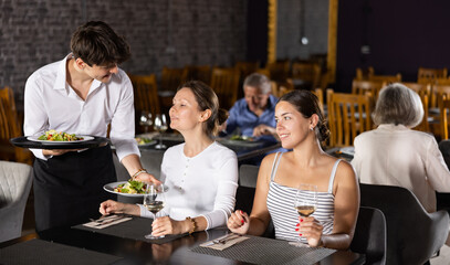 Young male waiter in uniform serves ordered dish to two women in restaurant..