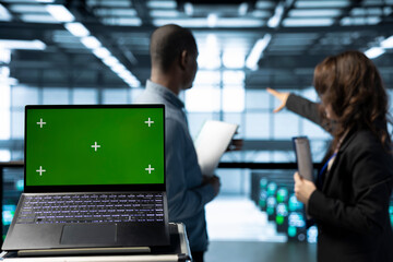 Green screen laptop in data center with programmers in blurry background employing machine learning models. Close up of chroma key notebook used by teamworking server room IT experts