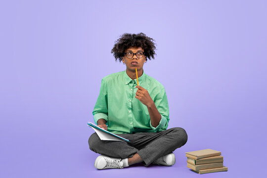 Thoughtful black guy student in glasses taking notes, holding notepad and pen, sitting over blue studio background, copy space. Cheerful teenager writing tasks or doing homework