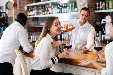 Young smiling bartender making a cool drink at the counter looking at two talking women