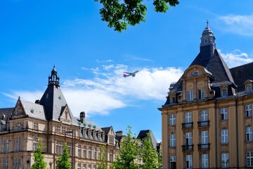 The historic buildings of Luxembourg City, Luxembourg showcase stunning architecture, with a plane flying overhead, blending modernity and history against a vibrant blue sky.