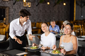 Friendly amiable young waiter talking to carefree laughing female friends gathered at dinner in restaurant, sitting at table and drinking wine